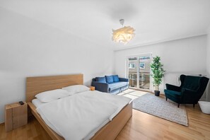 A modern bedroom with a wooden bed, a blue sofa, a dark armchair, and a potted plant. - Arbio I Flora Collection (Vienna)