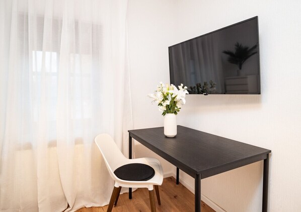 A small, modern room with a wooden floor, a white chair, a dark wooden table, a white vase with flowers, and a large flat-screen TV mounted on the wall. - Arbio | On Dorfstraße 82 (Nauen)