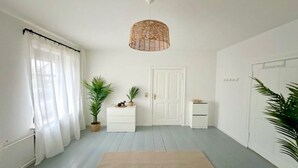 A room with a white door, a white dresser, a small white nightstand, a woven light fixture, and two potted plants.