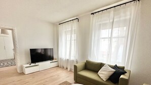 A modern living room with a flat-screen TV on a white stand, a green sofa with cushions, a white coffee table, and sheer curtains on the windows.