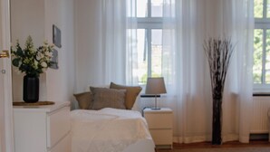 Quarto com piso de madeira, cama com cabeceira branca, mesa de cabeceira com abajur, um vaso com flores e janela com cortinas finas.