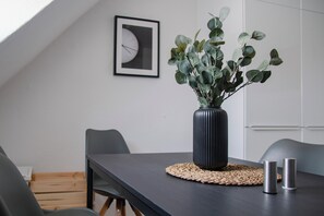 Un comedor moderno con una mesa oscura, un posavasos tejido, un florero negro alto con follaje verde y dos portavelas metálicos.