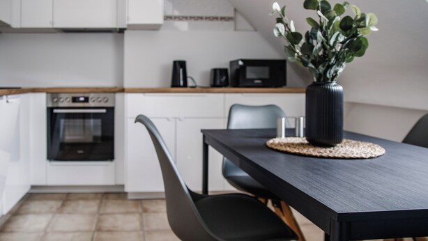 Cozinha moderna com armários brancos, bancada preta, micro-ondas e cafeteira. Há uma mesa de jantar com cadeiras e um vaso de flores.
