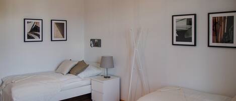 A bedroom with two beds, a bedside table with a lamp, and framed pictures on the wall.