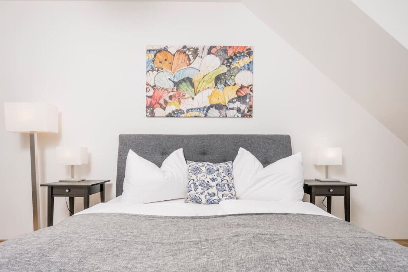 A bedroom with a grey headboard, white bedding, a colorful abstract painting, and two bedside tables with lamps.