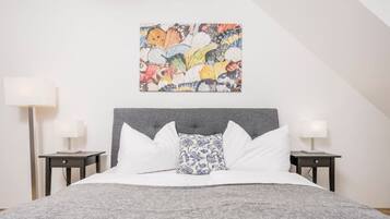 A bedroom with a grey headboard, white bedding, a colorful abstract painting, and two bedside tables with lamps.