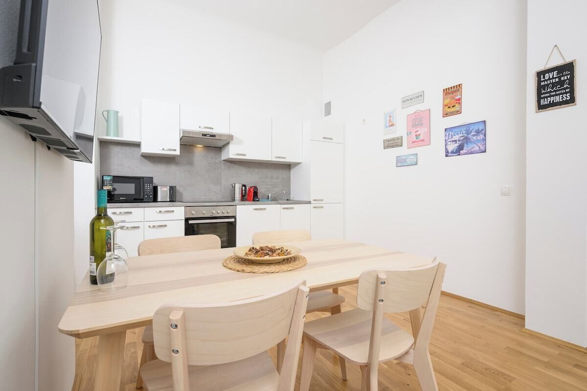Una cucina moderna con mobili bianchi, un forno a microonde e un tavolo da pranzo con sedie. Sono presenti decorazioni a parete e un cartello appeso.