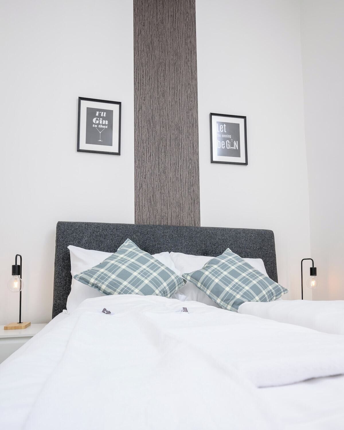 Una cama bien hecha con dos almohadas, un cabecero gris y dos mesitas de noche con lámparas. Hay dos citas enmarcadas en la pared.