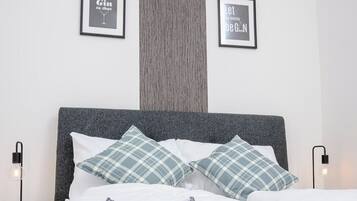 Una cama bien hecha con dos almohadas, un cabecero gris y dos mesitas de noche con lámparas. Hay dos citas enmarcadas en la pared.