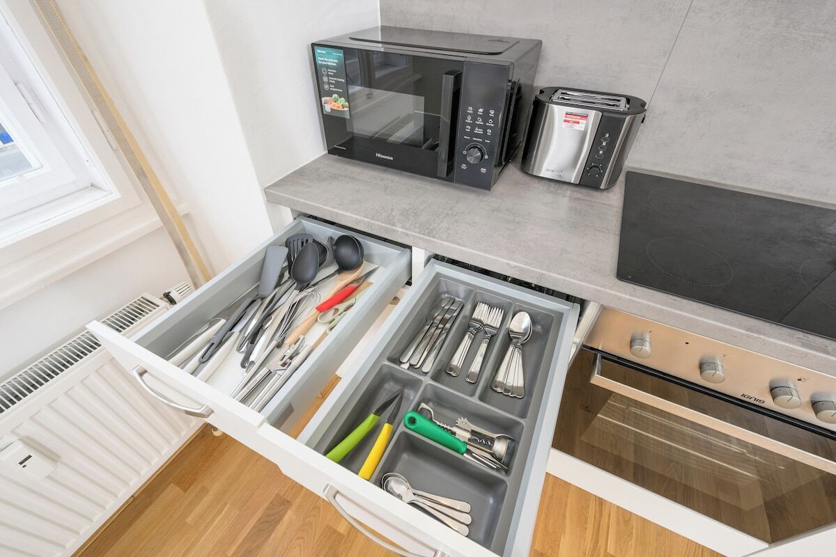 Een keuken met een magnetron, broodrooster en oven. Er is een lade met keukengerei en een raam boven de keuken.