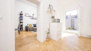 A modern living room with a herringbone floor, a grey sofa, a white cabinet, and a view of the outside through an open door. - Anna And Friends (Graz)
