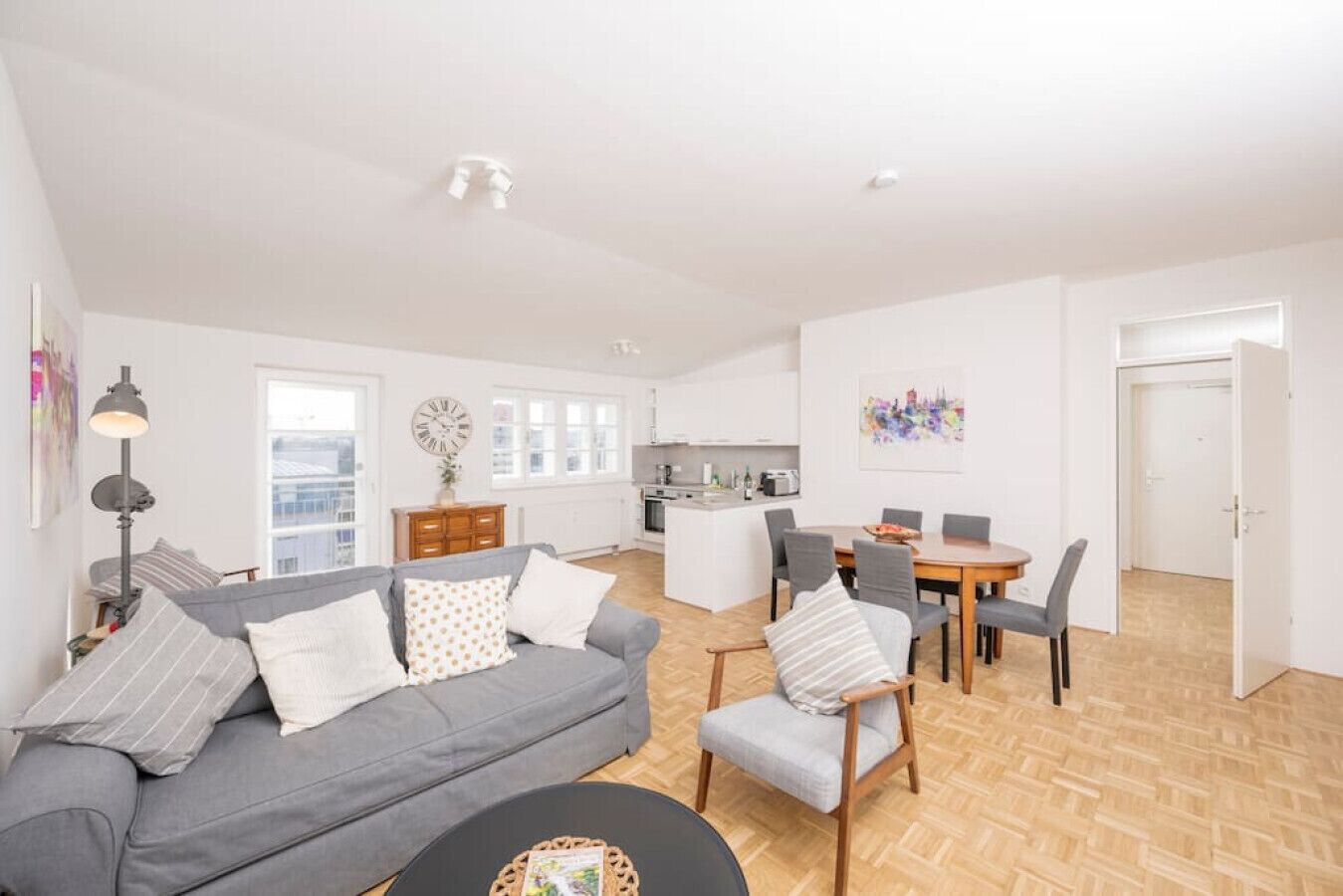 A modern apartment with a grey sofa, a dining table with chairs, a kitchen area, and a large window.