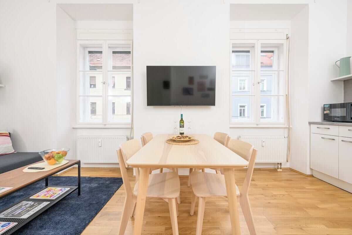 A modern living room with a wooden dining table, six chairs, a flat-screen TV mounted on the wall, and a kitchen area with a microwave.