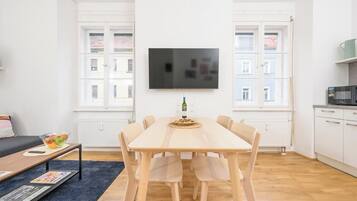 A modern living room with a wooden dining table, six chairs, a flat-screen TV mounted on the wall, and a kitchen area with a microwave.