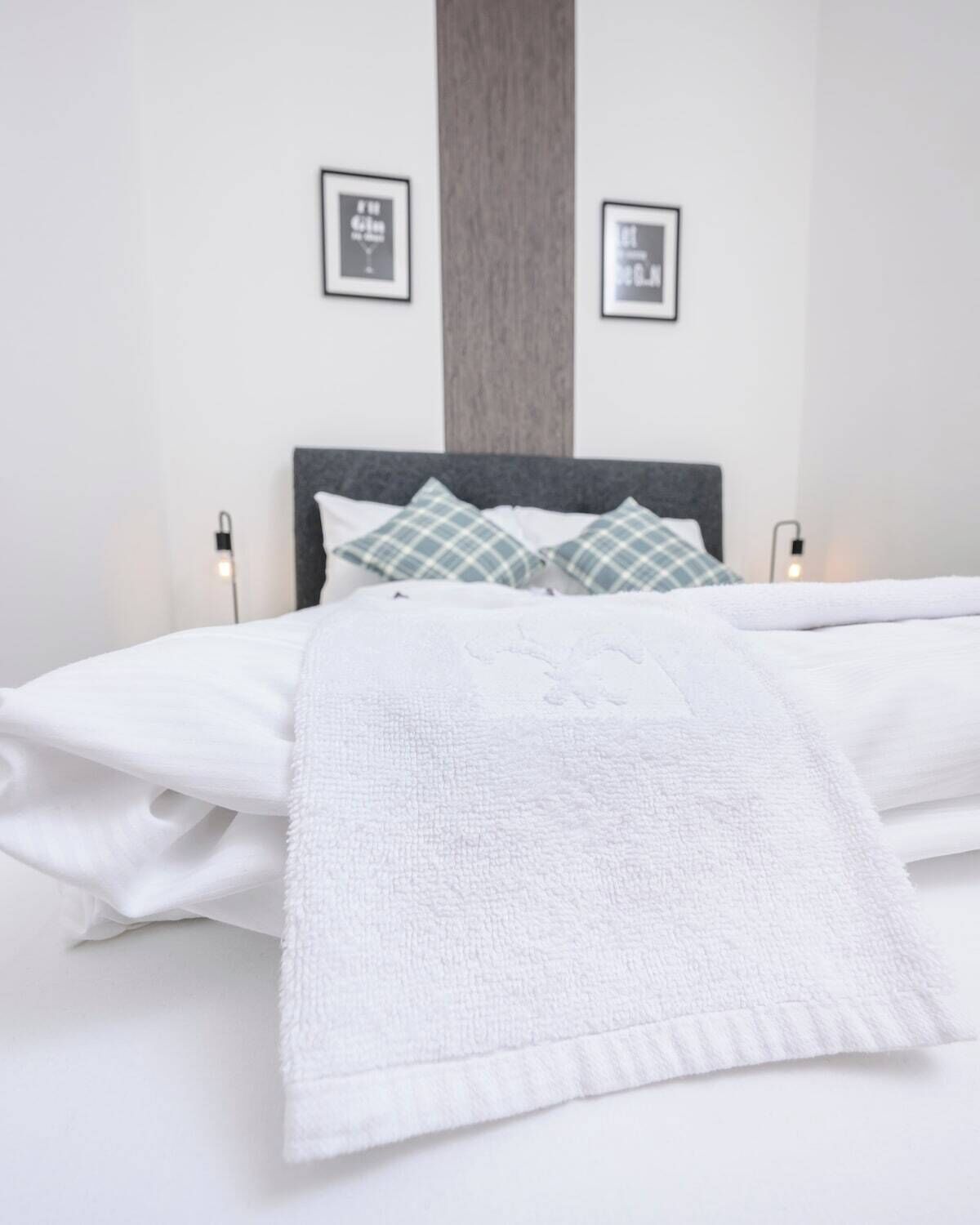 A neatly made bed with white linens and a white throw blanket. There are two pillows with a checkered pattern, and the bed is flanked by two bedside lamps.
