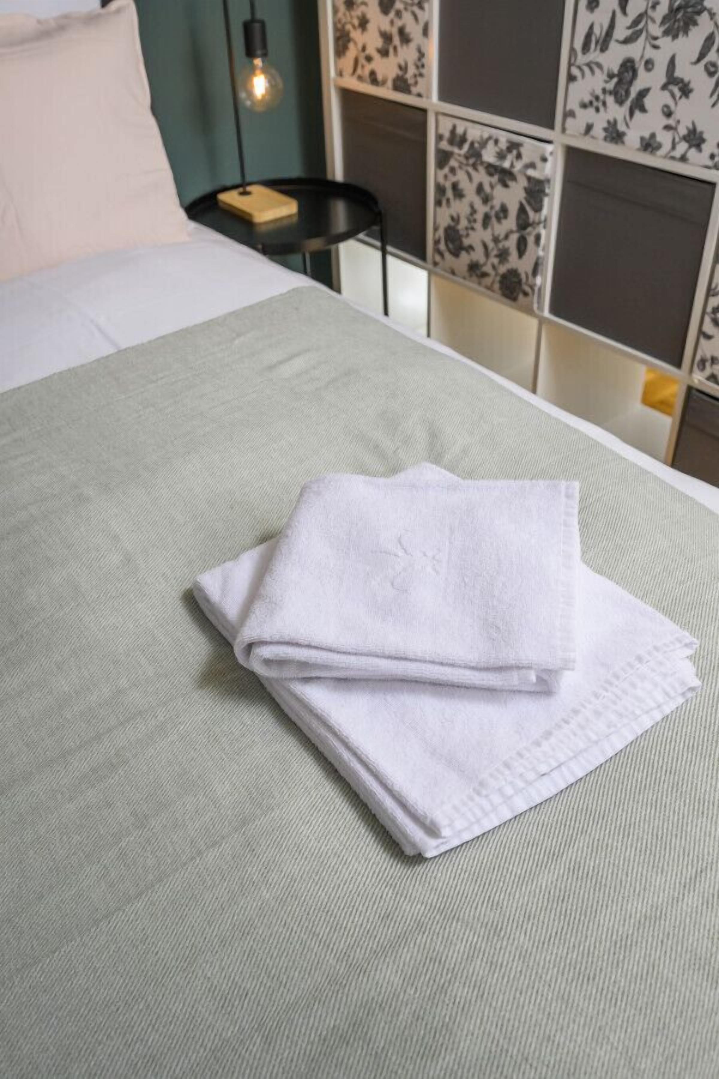 A neatly made bed with a folded white towel on a green bedspread, a black nightstand with a lamp, and a floral-patterned headboard.
