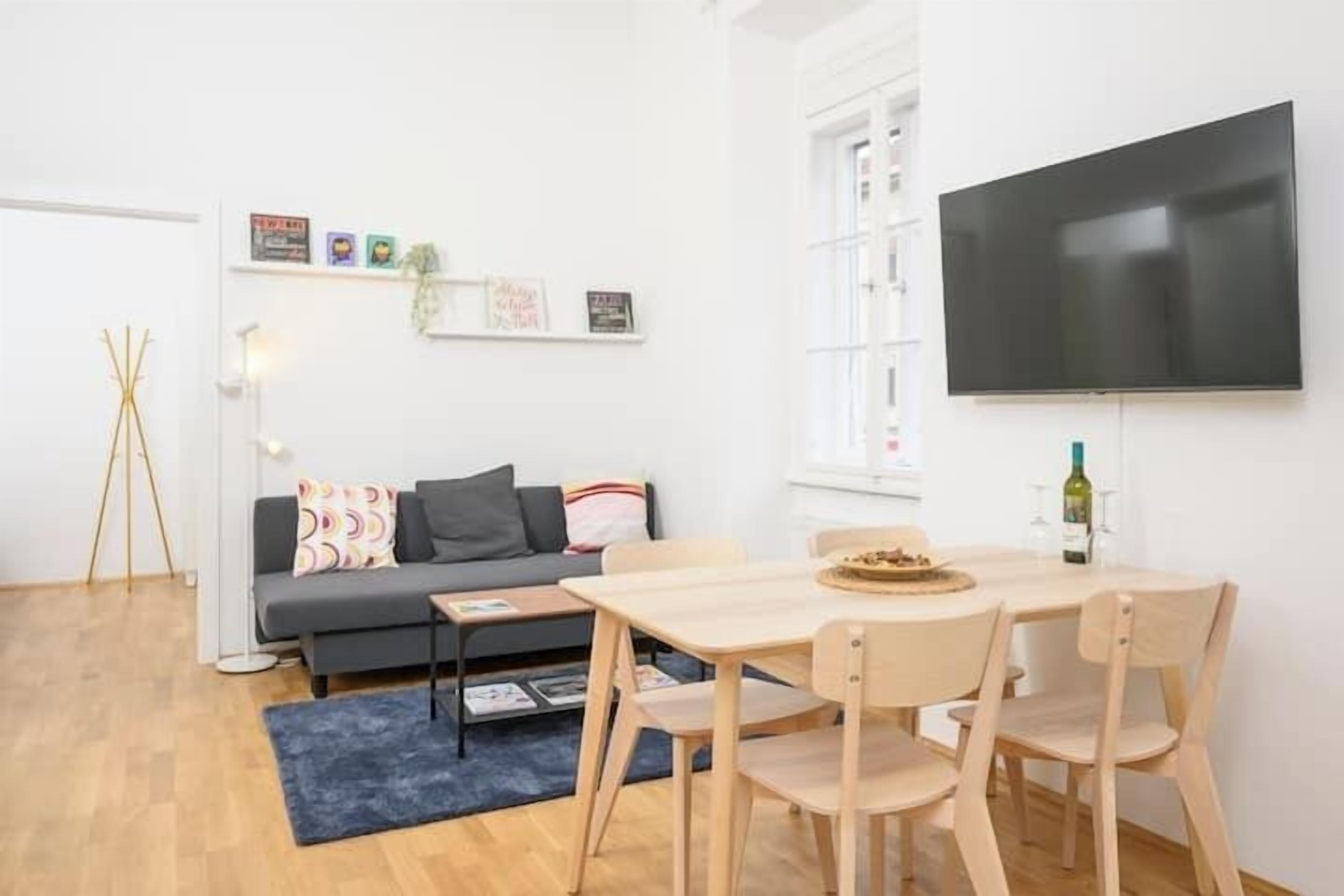 Un salon moderne comprenant un canapé, une table à manger avec des chaises, un téléviseur à écran plat fixé au mur et une petite table d’appoint avec une bouteille et des verres.