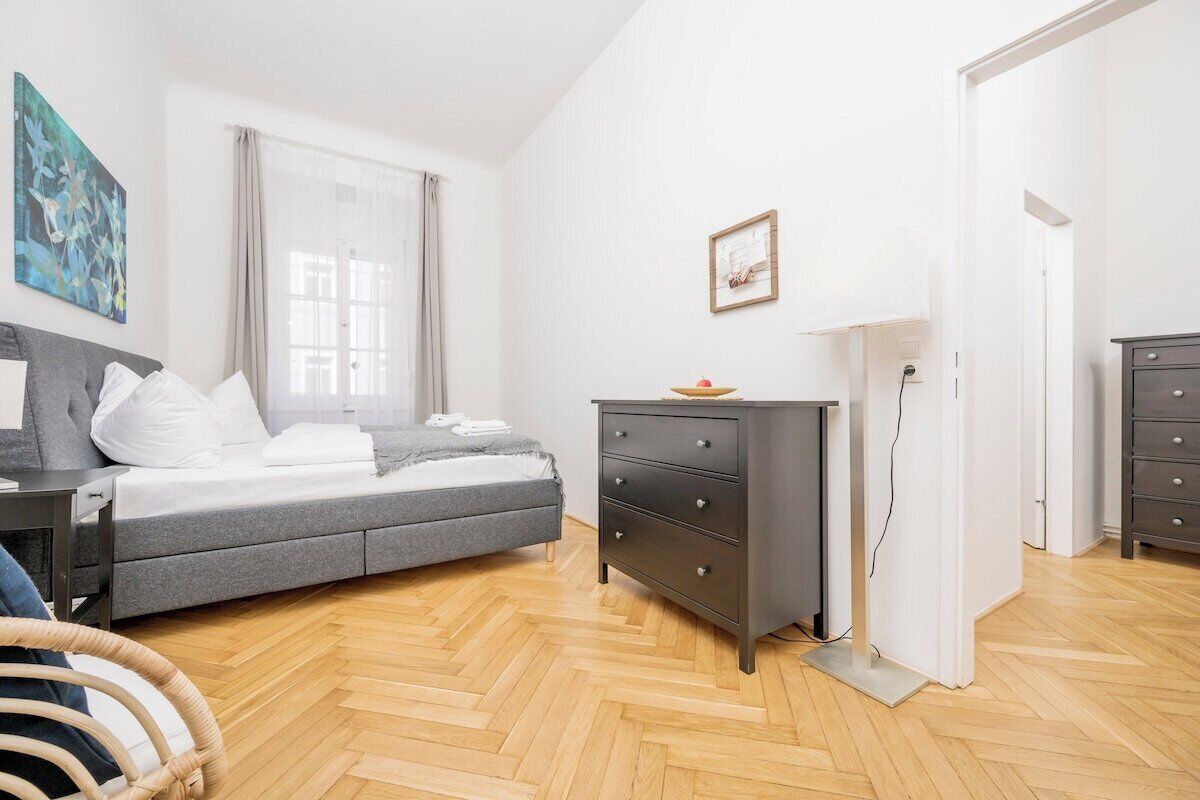 Une chambre moderne avec un canap é-lit gris, une commode sombre et un parquet en bois disposé en chevrons.