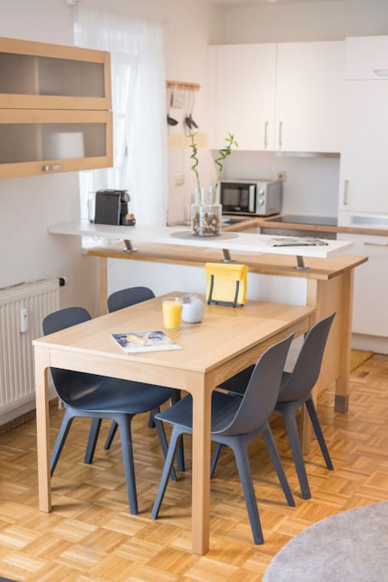 A modern kitchen with white cabinets, a microwave, and a toaster. There is a wooden dining table with four chairs, and a small plant on the counter.