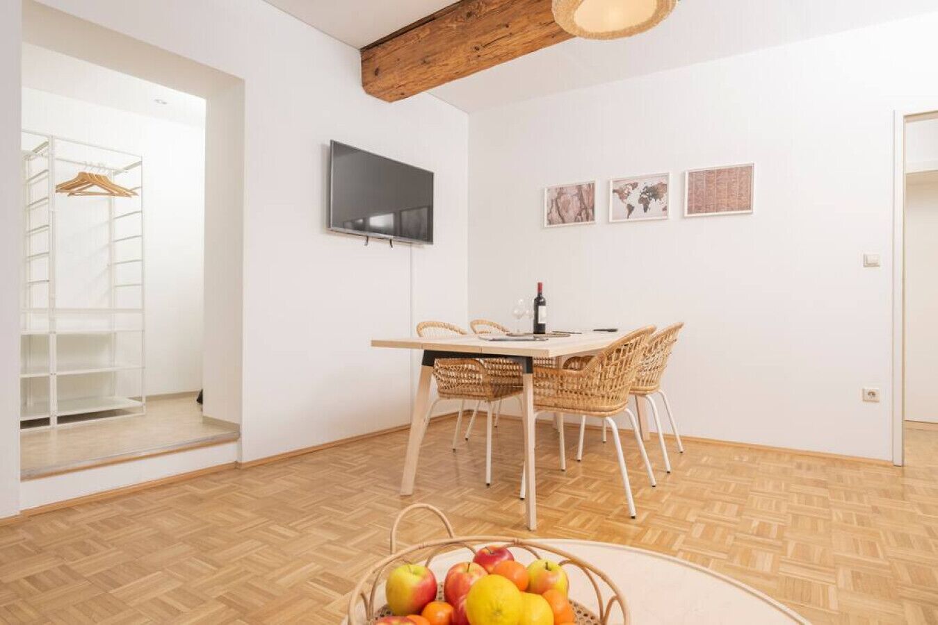 A modern living room with a wooden ceiling, a TV mounted on the wall, a dining table with wicker chairs, and a fruit basket on the floor.