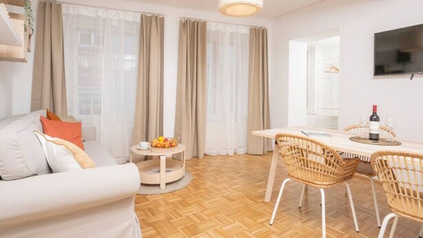 A modern living room with a sofa, dining table, wicker chairs, a small round table with fruit, and a TV on the wall. - Zum Grazer Uhrturm (Graz)