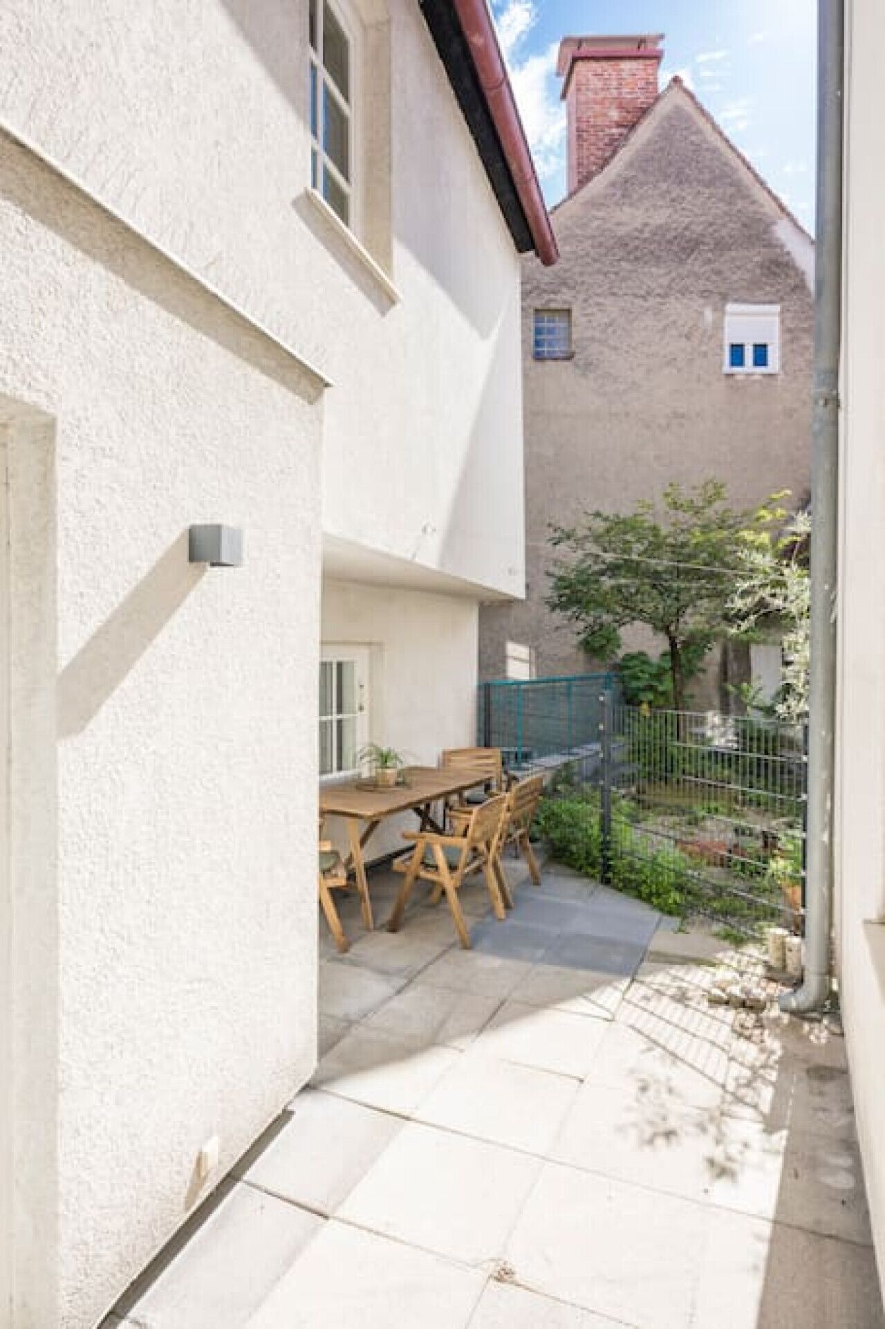 A narrow outdoor patio with a wooden table and chairs, a small garden area, and a metal fence.