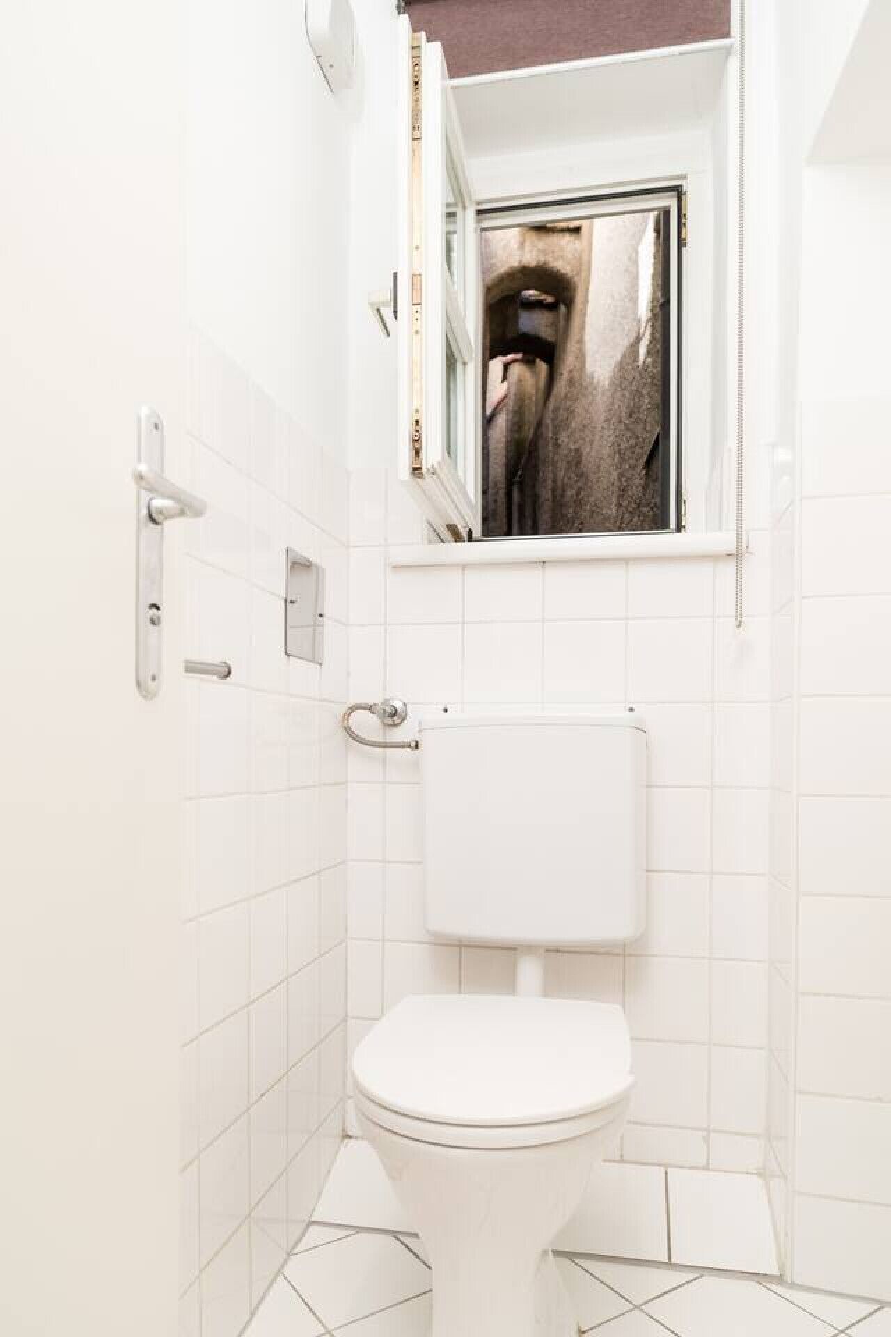Un baño pequeño y blanco con un inodoro, un espejo sobre el inodoro y una ventana.