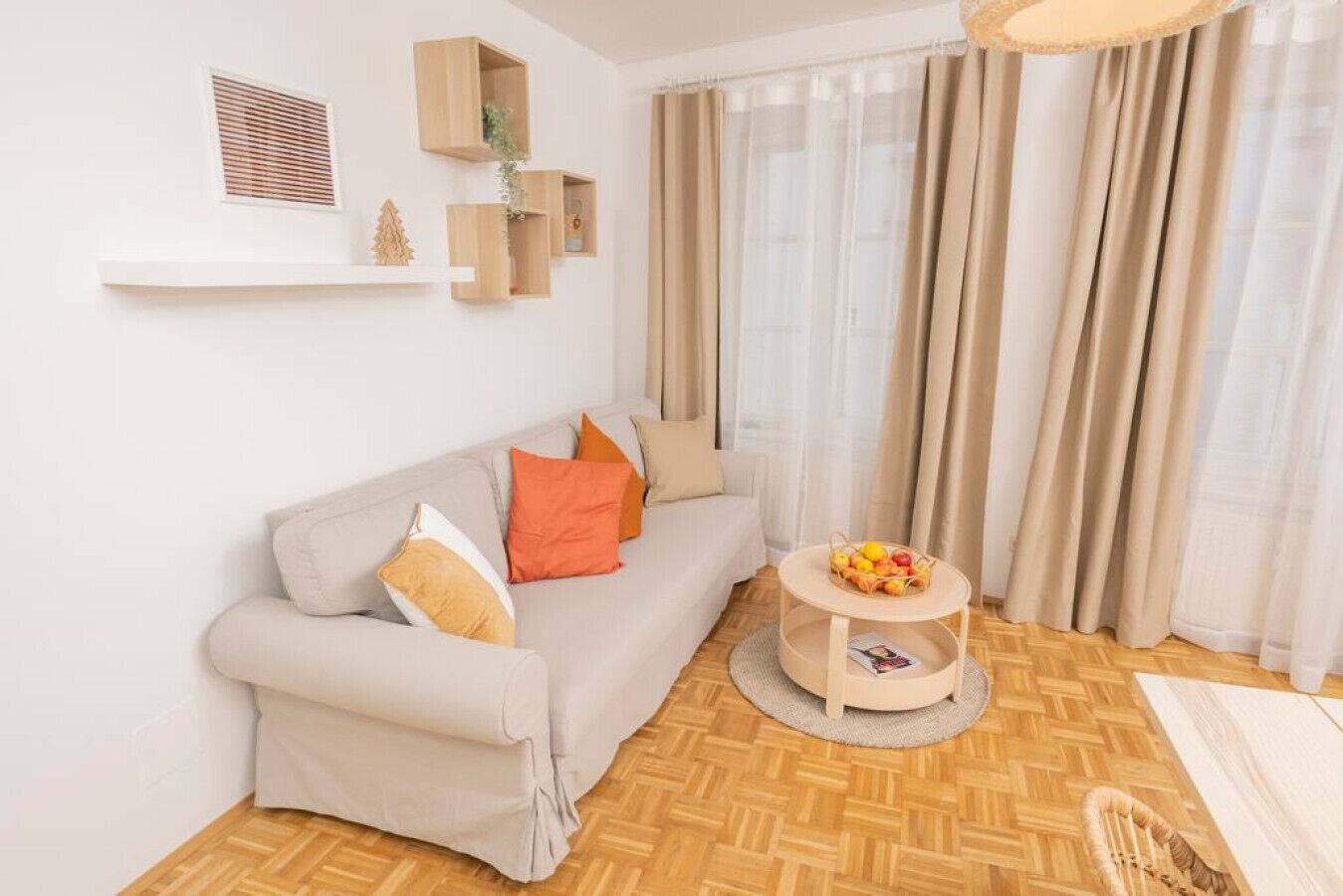 Ein Wohnzimmer mit einer Couch, Kissen, einem runden Holztisch mit Früchten und einem Fenster mit Vorhängen.