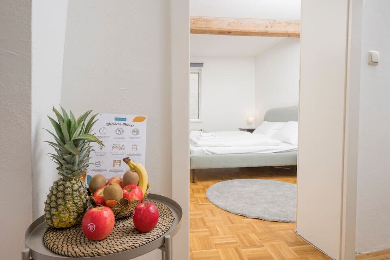 A room with a bed, a small table with fruits, and a welcome sign.