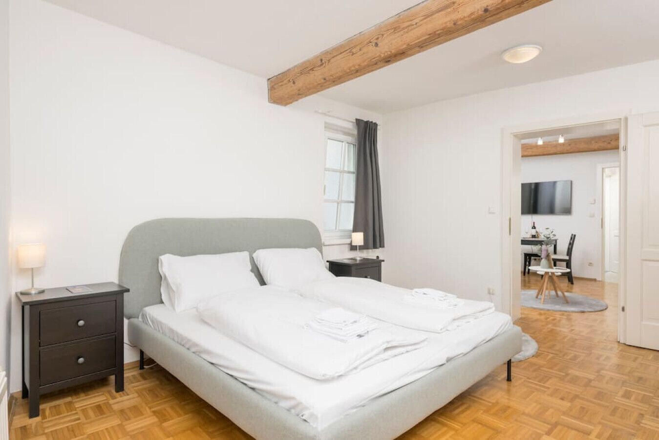 A bedroom with a bed, bedside tables, a lamp, a window with curtains, and a wooden ceiling beam.