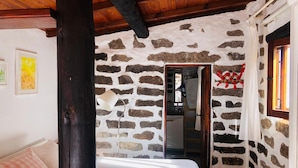 A cozy room with a stone wall, a bed with a white sheet, a wooden ceiling, and a window with curtains.