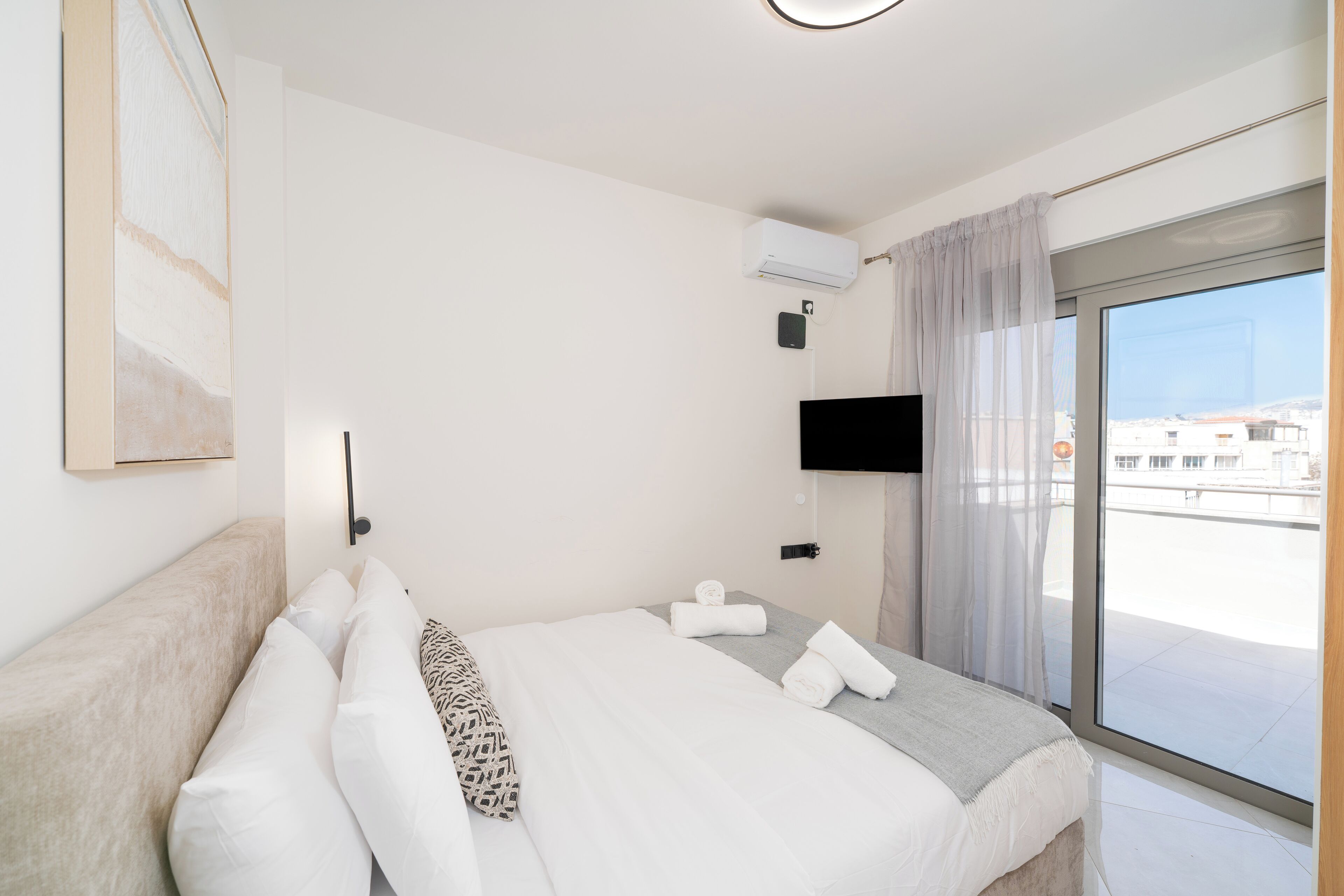 A modern hotel room with a large bed, a flat-screen TV mounted on the wall, and a large window offering a city view.