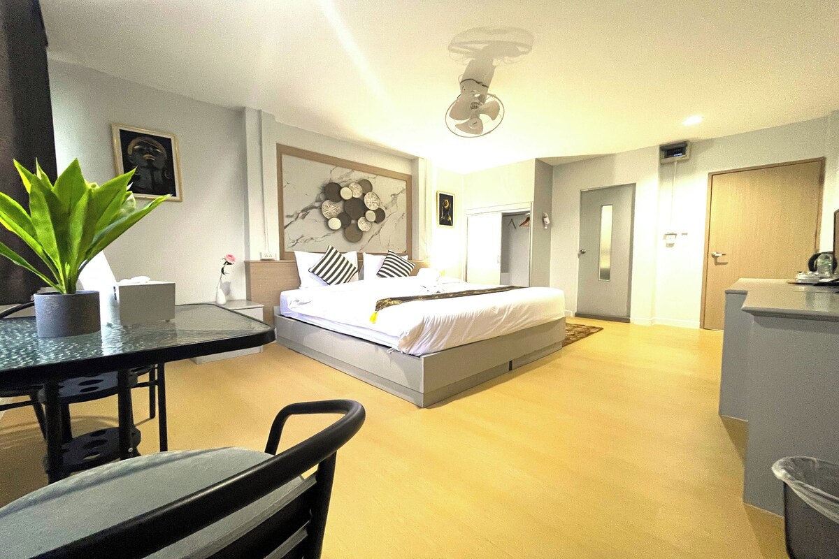 A modern hotel room with a bed, a desk, a chair, a potted plant, and a ceiling fan.