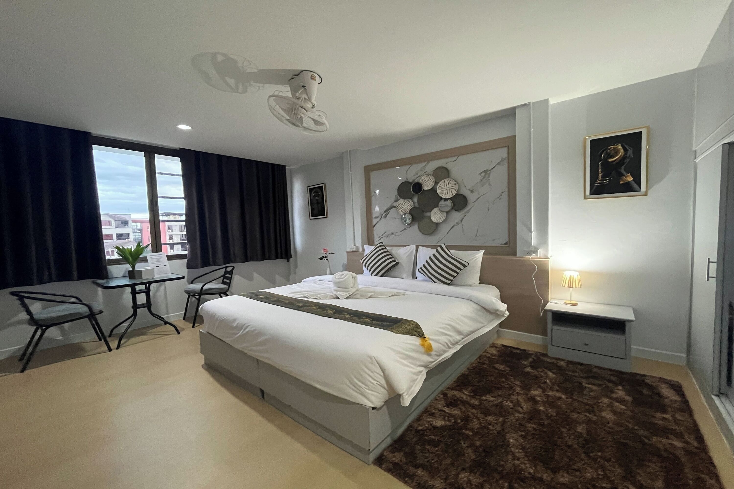 A modern hotel room with a large bed, a desk, and a chair. There is a window with curtains, a wall-mounted artwork, and a small table with a lamp.