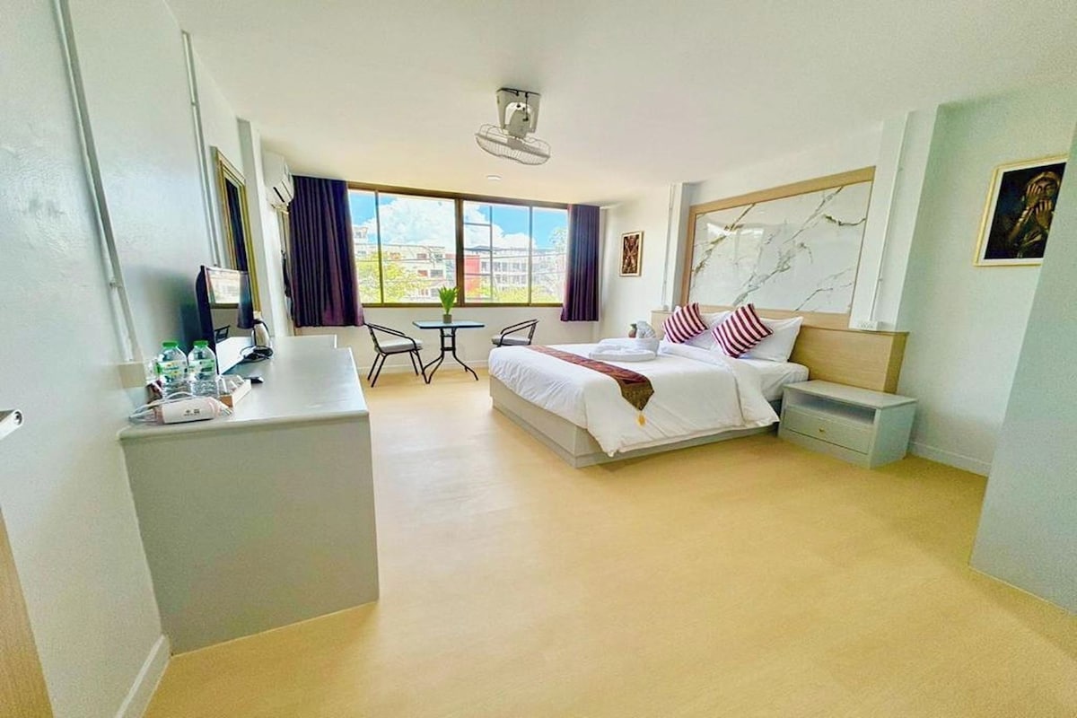 A hotel room with a bed, bedside table, a small desk with a chair, a window with curtains, and a view of buildings outside.