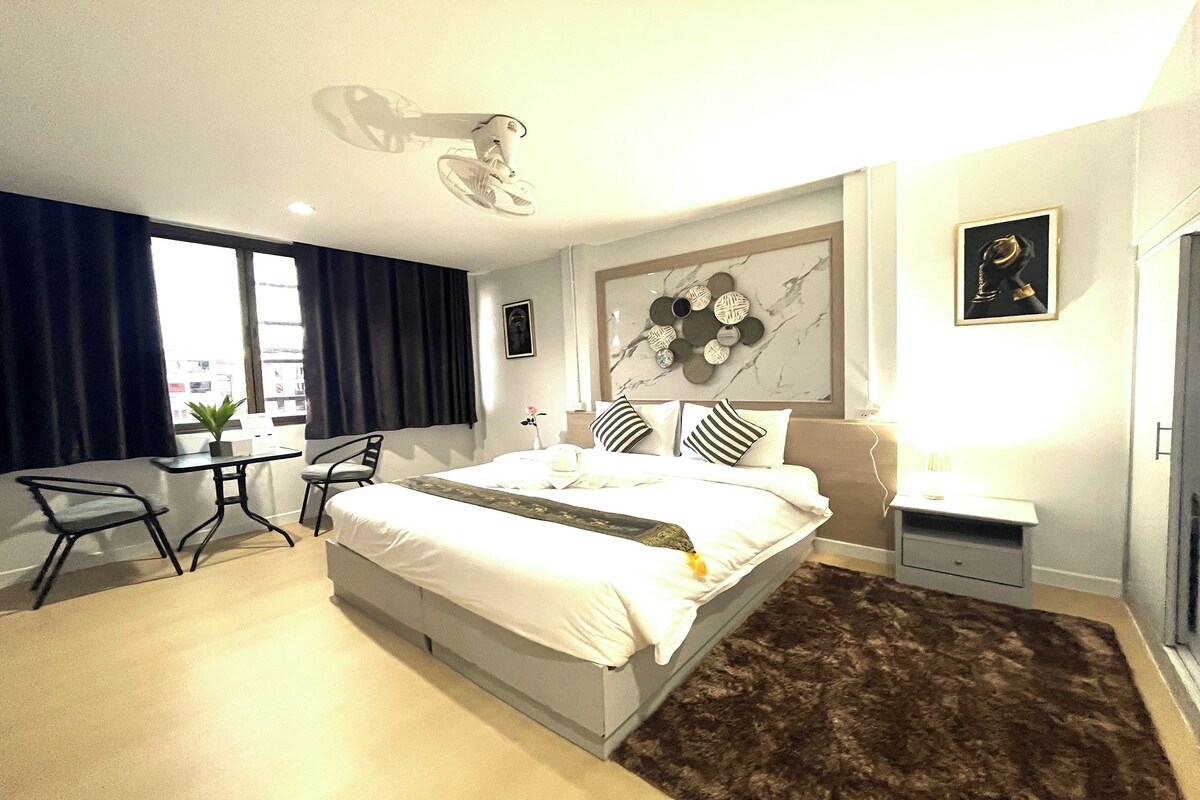 Een moderne hotelkamer met een groot bed, een bureau en een stoel. Er is een raam met gordijnen, een wandlamp en een klein tafeltje met een plantje.
