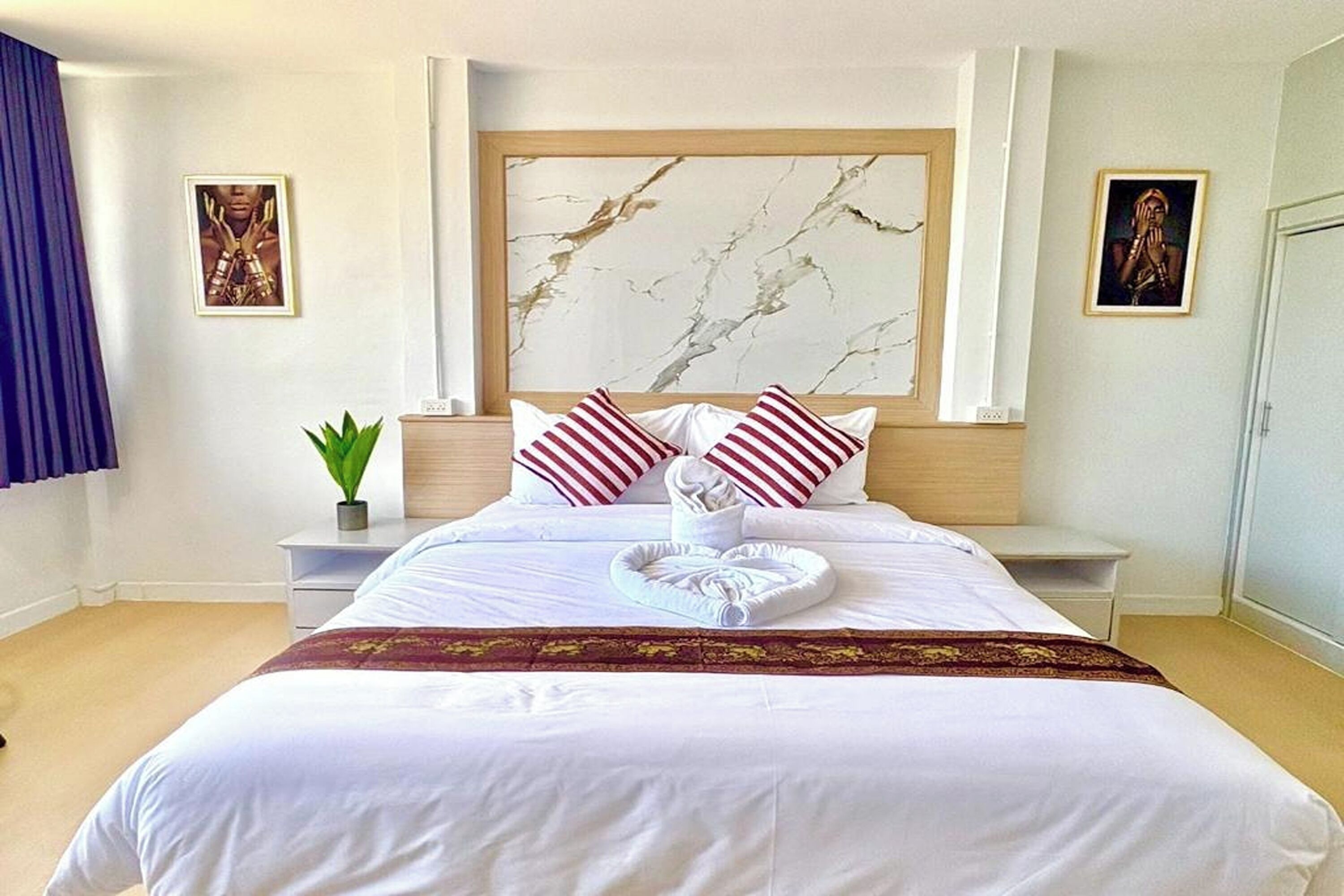 A hotel room with a large bed, white bedding, red and white striped pillows, a marble headboard, a potted plant, and artwork on the walls.