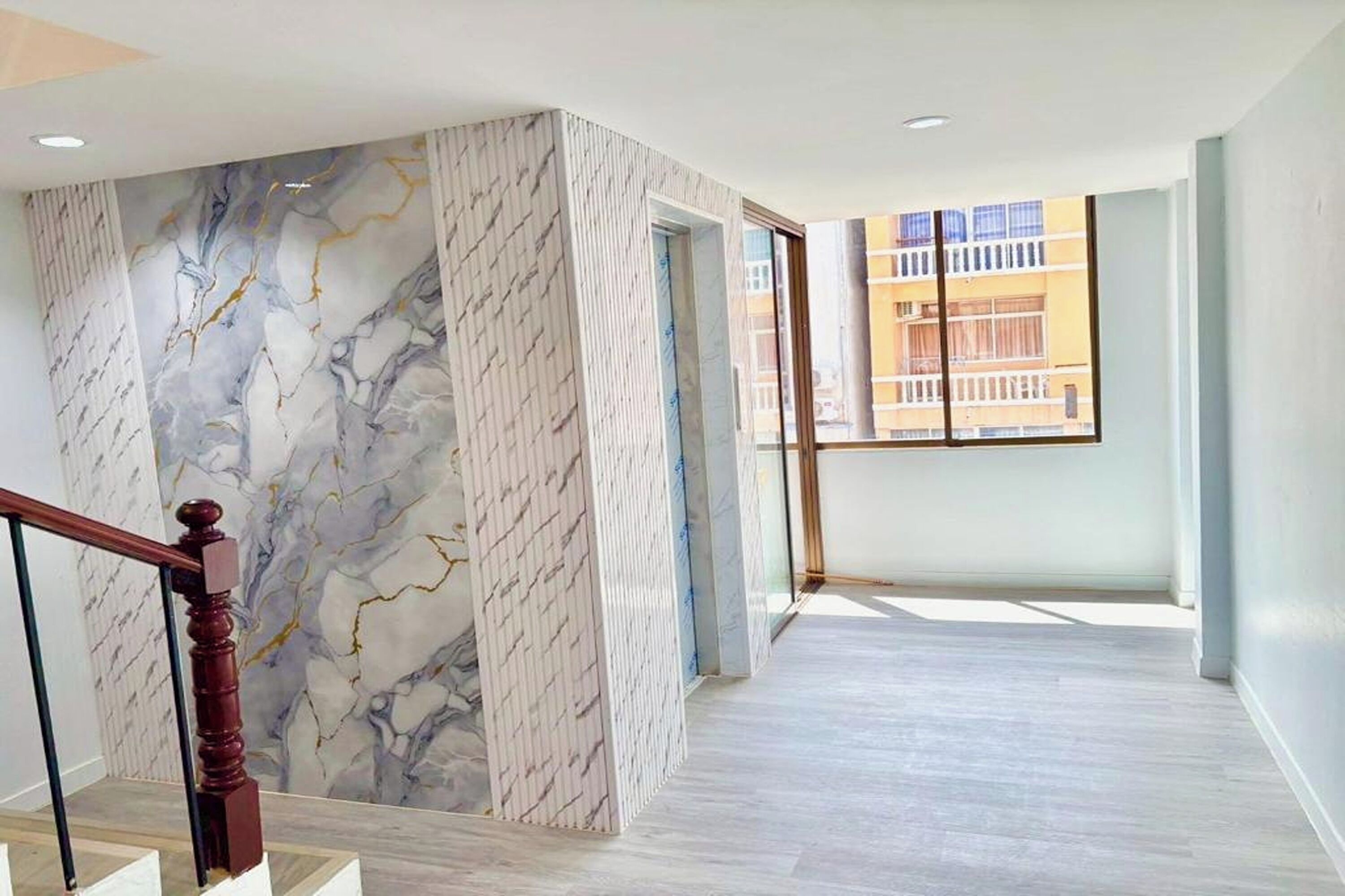 A spacious interior with a marble wall, a wooden staircase, and a large window offering a view of buildings outside.