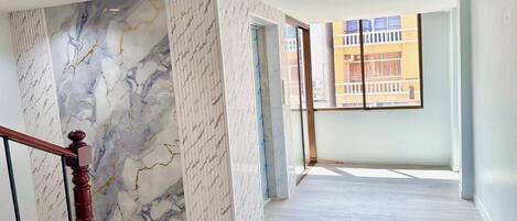 A spacious interior with a marble wall, a wooden staircase, and a large window offering a view of buildings outside.
