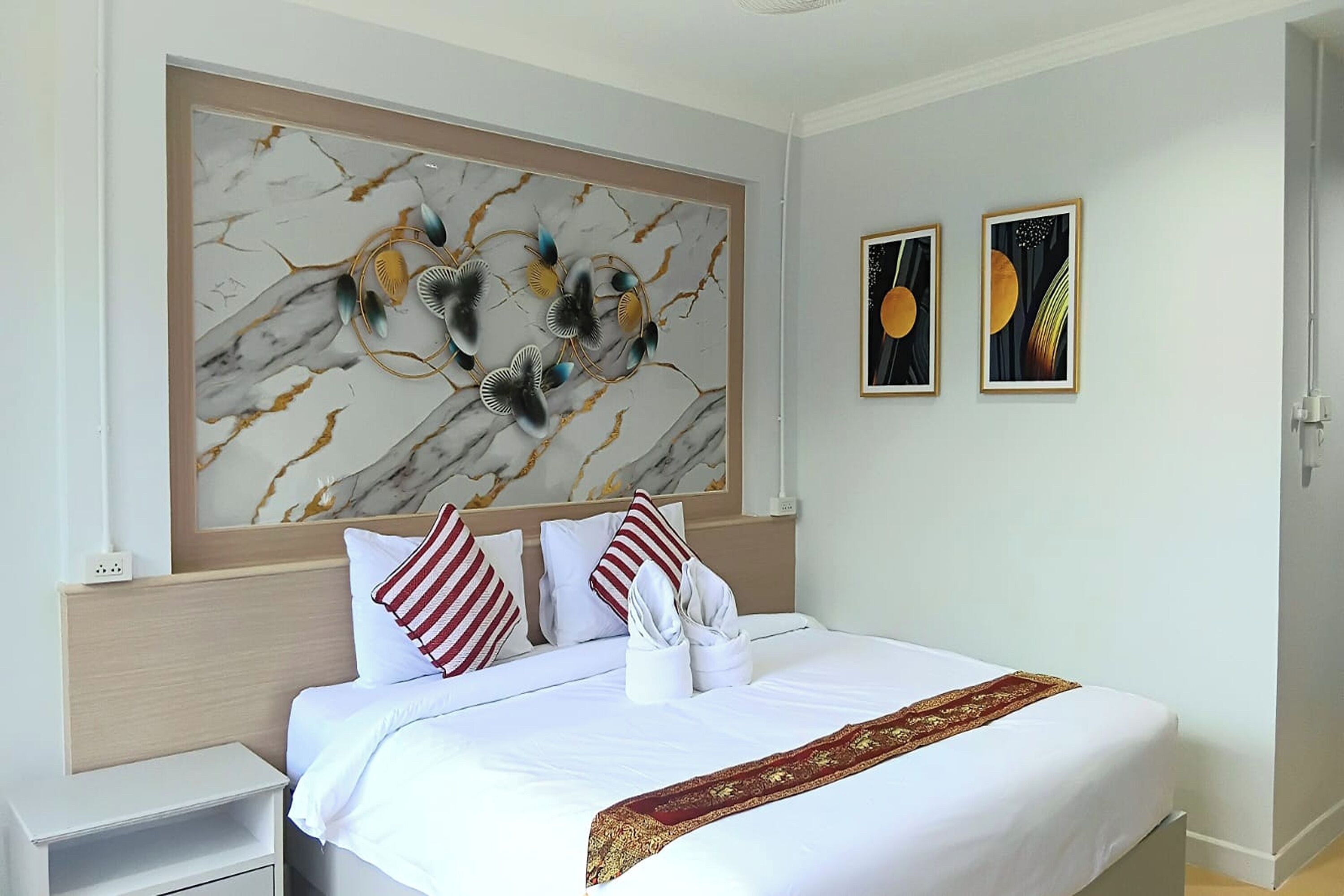 A hotel room with a marble-patterned wall art, a neatly made bed with white linens and a red-striped pillow, and two framed artworks on the wall.