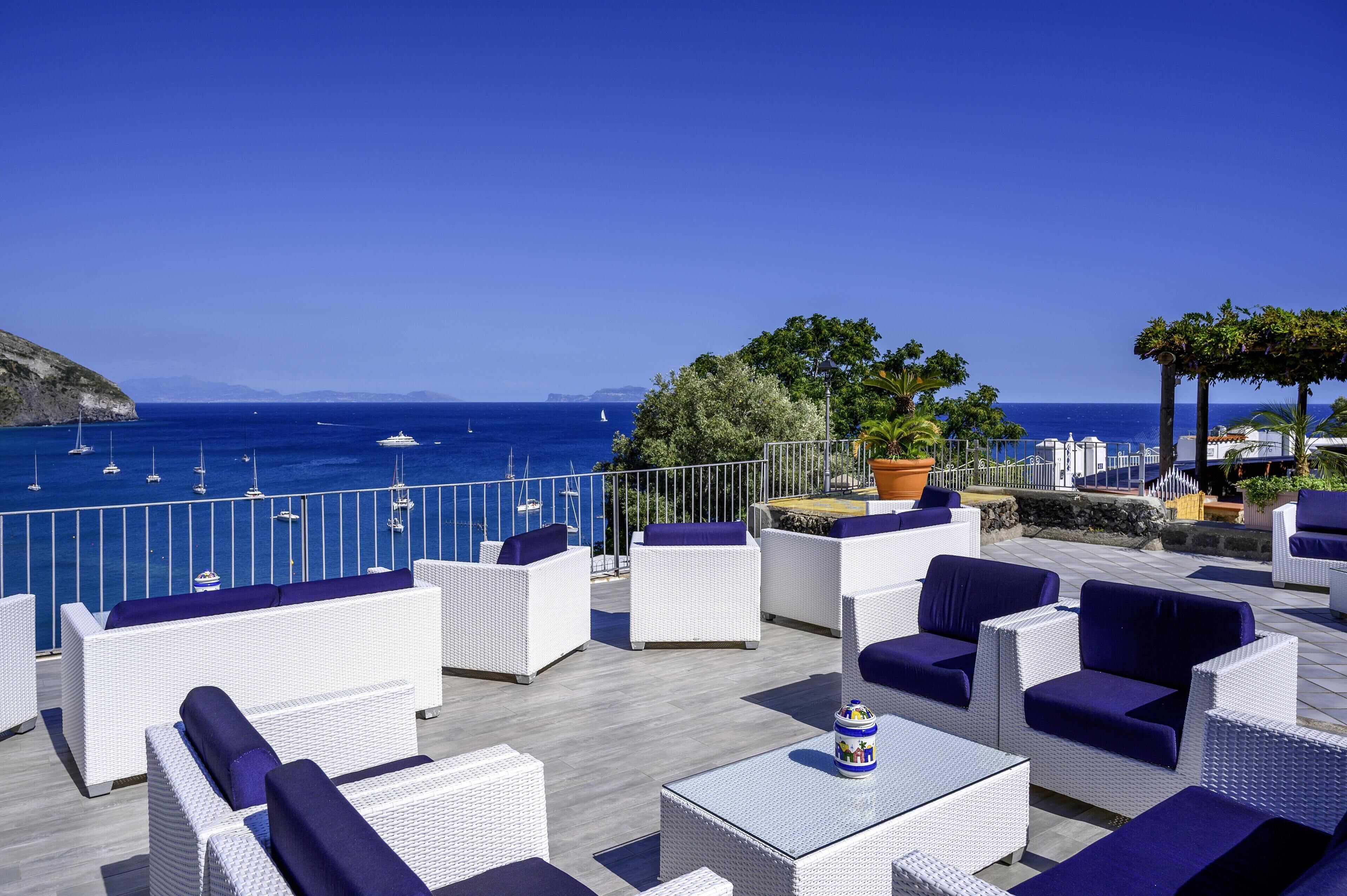A terrace with white wicker furniture and blue cushions, overlooking the sea with boats and a clear sky.