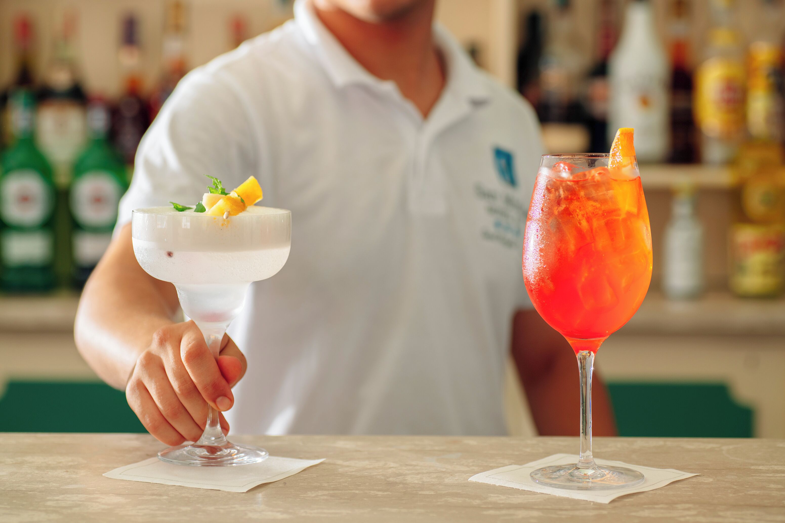 A bartender holding two drinks, one in a coupe glass and the other in a highball glass, with a blurred background of bottles.