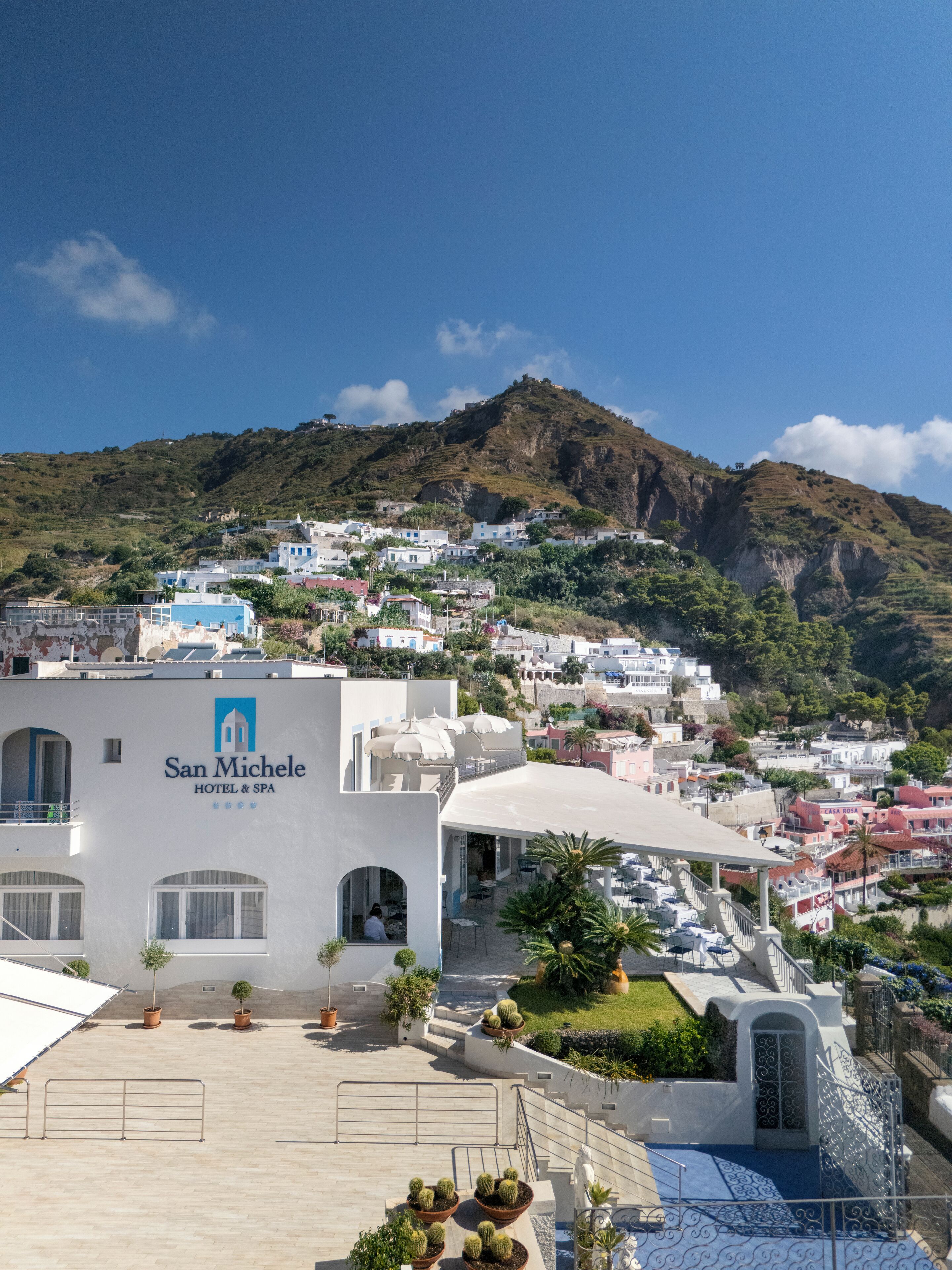A hotel named San Michele with a rooftop terrace, a pool, and a view of the surrounding hillside town.