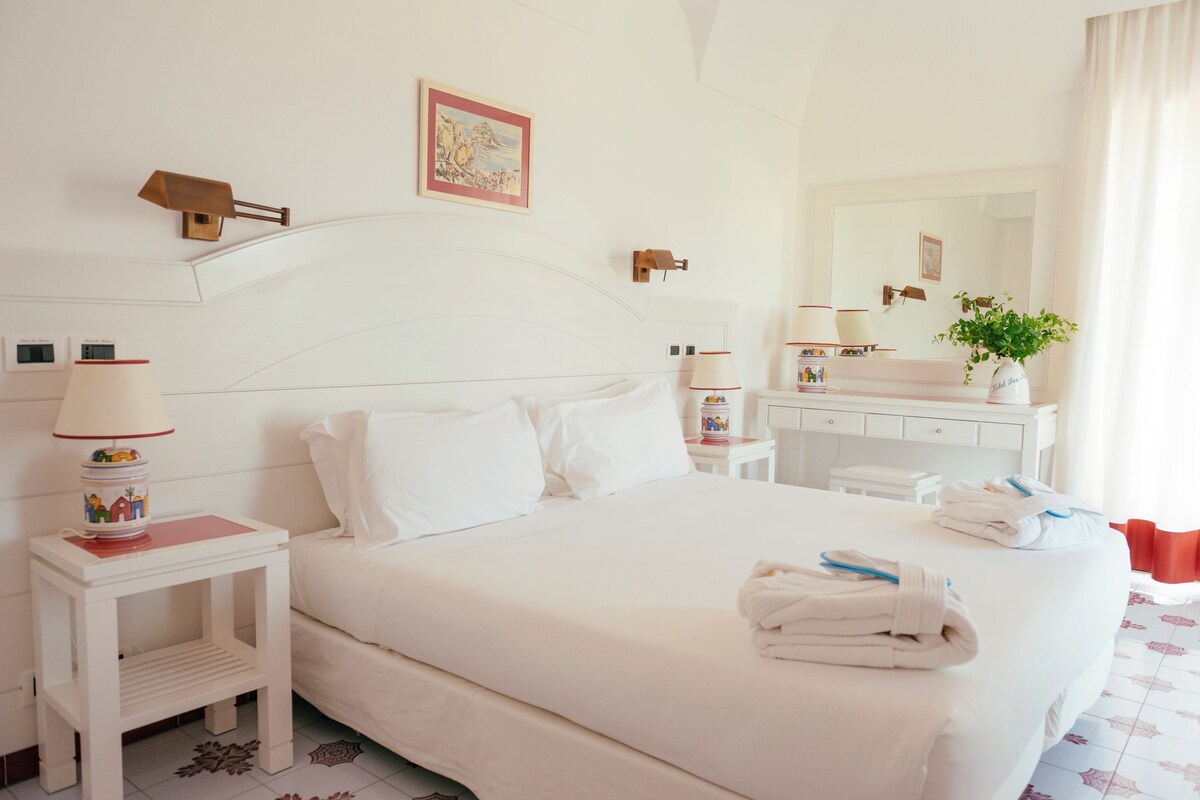 A neatly made bed with white linens, a bedside table with a lamp, a small plant, and a mirror above the dresser.