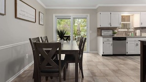 Un coin repas avec une table et des chaises, une cuisine avec des meubles blancs et, derrière une porte vitrée, une vue sur un patio extérieur.