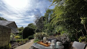 A patio with a round table and two chairs, a tray with cups and a jar, and a garden with various plants and a pathway.
