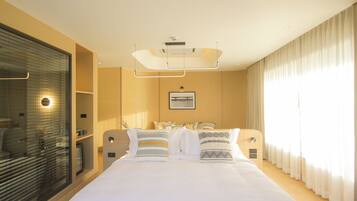 A modern hotel room with a large bed, a bedside table, a mirror, a window with curtains, and a wall-mounted artwork.