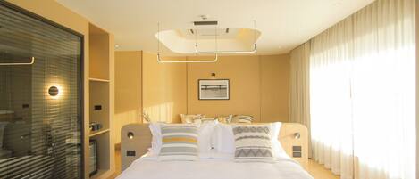 A modern hotel room with a large bed, a bedside table, a mirror, a window with curtains, and a wall-mounted artwork.