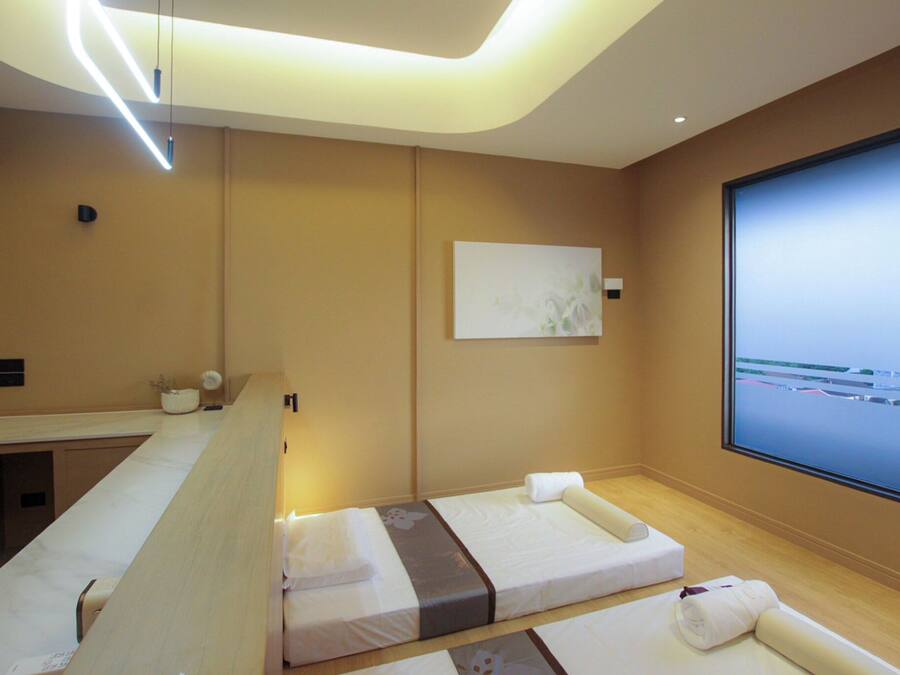 A modern hotel room with a large window, a massage table, a small bedside table, and a wall-mounted artwork.