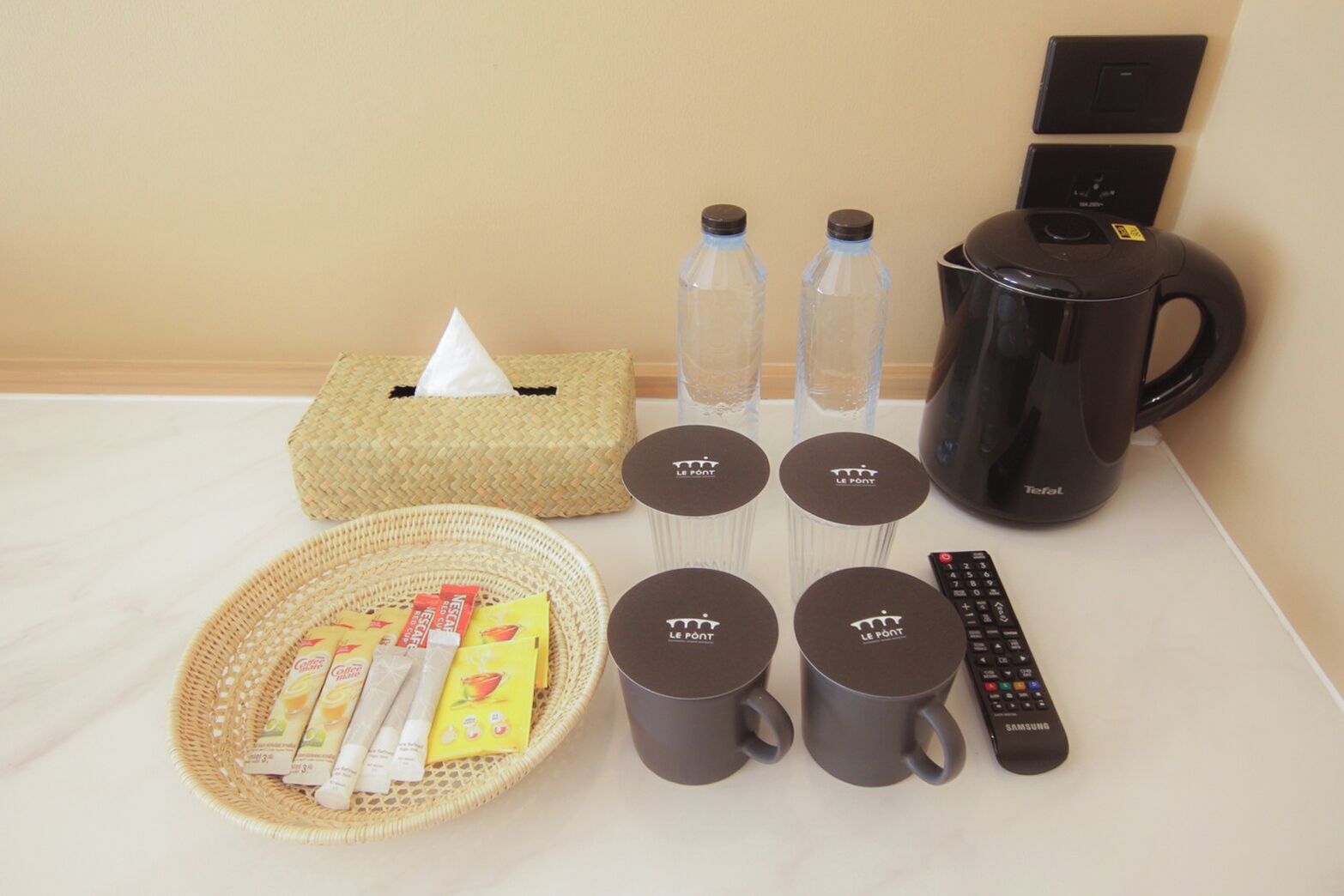 A hotel room with a coffee maker, water bottles, a tissue box, a basket with toiletries, and a remote control.